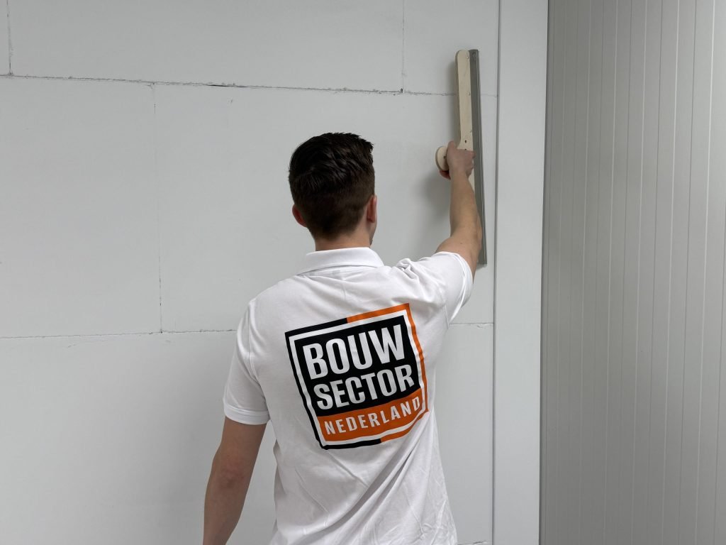 Close-up van een stukadoor van Bouwsector Nederland die een brede spaan boven zijn hoofd tegen de wand houdt