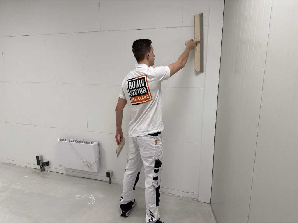 Stukadoor in wit tenue staat naast een elektrische kachel en zet een lange spaan hoog tegen de muur
