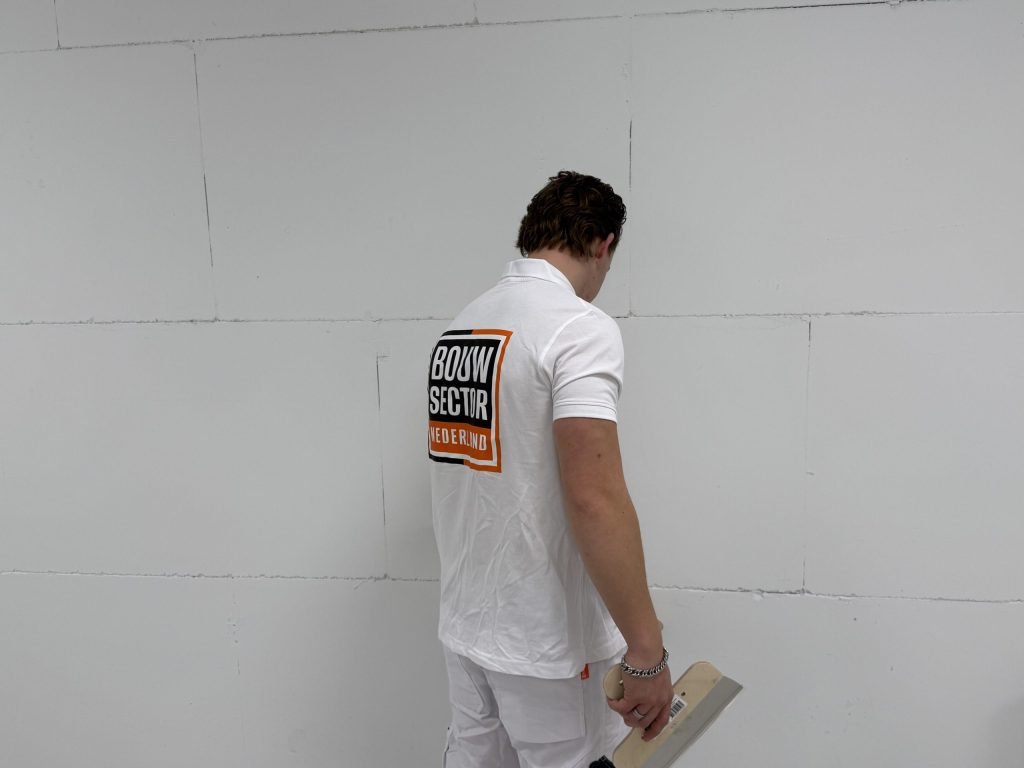 Stukadoor van Bouwsector Nederland bekijkt van dichtbij een kale wand met naden, een plakspaan in de hand